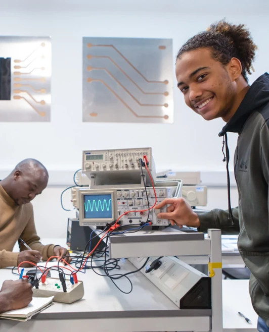 Three students engaged in an electronics lab experiment. One student writes notes while another adjusts an oscilloscope, with circuit-themed wall panels behind them. Three students engaged in an electronics lab experiment. One student writes notes while another adjusts an oscilloscope, with circuit-themed wall panels behind them.